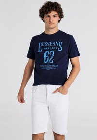 Camiseta de algodón azul marino con gráfico "Lois Jeans Legendary 62" en azul claro, combinada con shorts de mezclilla blancos. Ajuste casual.