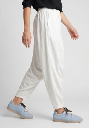 Persoon die loopt met losse witte broek met elastische tailleband en lichtblauwe sneakers met contrasterende veters op een effen achtergrond.