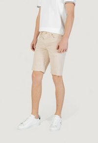 Pantaloni corti beige con frontale piatto, due tasche laterali e un tessuto leggermente texturizzato. Indossati con sneakers bianche e una polo bianca.