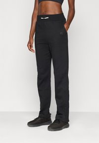 Zwarte trainingsbroek met een geribbelde tailleband, zijzakken en textuurdetails langs de benen. Gepaard met zwarte sportschoenen.