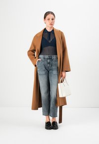 Donna con un cappotto marrone sopra una blusa trasparente blu navy, jeans grigi a vita alta, mocassini neri e una borsa bianca in mano, su sfondo bianco.