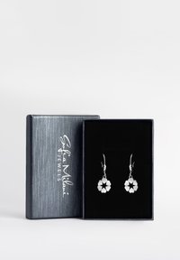 Silver blommformade örhängen i en svart sammetfodrad låda, med en stilren silverexteriör som har en vit logotyp.