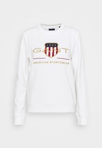 Bílá mikina s dlouhými rukávy, zdobená vyšívaným štítovým logem v červené a zlaté barvě a nápisem „AMERICAN SPORTSWEAR“ pod ním.