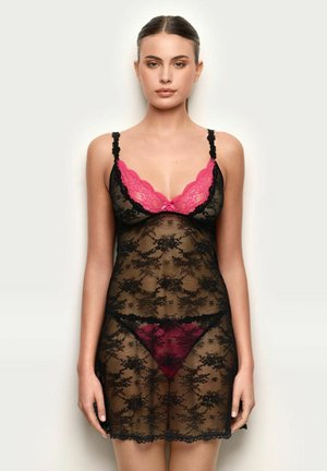 Mujer de cabello oscuro recogido, vestida con un vestido de lencería de encaje negro transparente y ropa interior de encaje rosa y rojo oscuro, de pie contra un fondo liso.