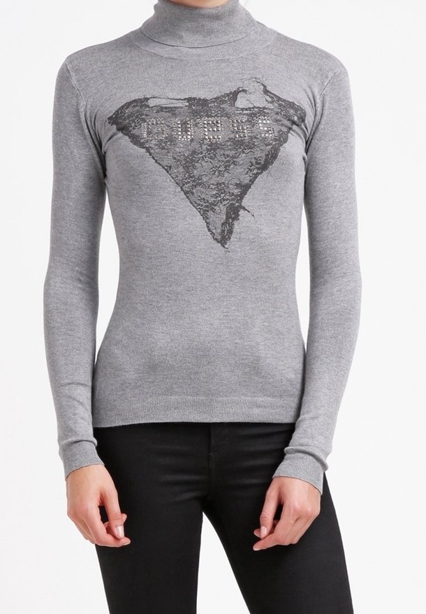 Personne portant un pull gris à col roulé à manches longues avec un triangle en dentelle noire et un logo pailleté sur la poitrine, assorti à un pantalon noir.
