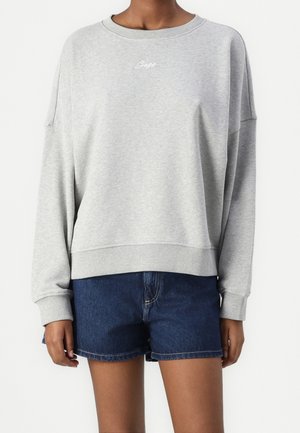 Osoba ubrana w jasnoszarą bluzę z długim rękawem z napisem „Boss” na piersi oraz ciemnoniebieskie dżinsowe szorty, stojąca na białym tle.