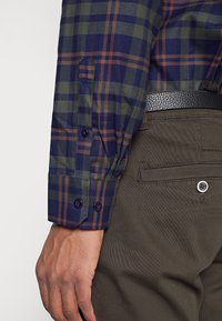 Camicia a maniche lunghe a quadri con un motivo a scacchi blu navy, verde e arancione. Il tessuto ha una texture liscia, con polsini a bottoni e una cintura scura.