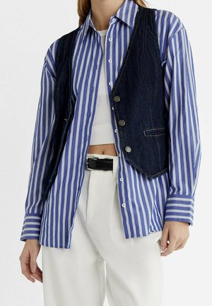 Frau trägt ein blau-weiß gestreiftes, geknöpftes Hemd, eine dunkelblaue Jeansweste mit Knöpfen, ein weißes kurzes Top, weiße Hose und einen schwarzen Gürtel.