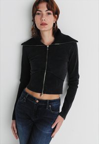 Črna skrajšana jakna z širokim ovratnikom in zadrgo, izdelana iz mehkega materiala. V kombinaciji s temnimi jeans hlačami z sprednjimi žepi.