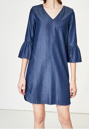 Nainen, joka pukeutunut polvipituiseen, farkunsiniseen mekkoon, jossa on V-kaula-aukko ja kolmen neljänneksen liehuhihat, yksinkertaista taustaa vasten.