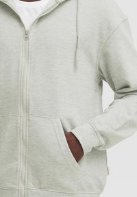 Felpa grigia chiara con zip, realizzata in tessuto morbido, dotata di cappuccio con coulisse, tasche frontali e polsini a coste. Leggera texture su tutta la superficie.