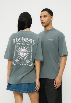 Zwei Modelle tragen ausgewaschene grüne, übergroße T-Shirts; eines zeigt die Rückseite mit einem Grafikdesign von Sonne und Mond und dem Text „ALCHEMY of the Soul“, das andere blickt nach vorne.