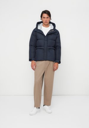 Marineblauwe pufferjack met een capuchon, voorzien van een ritssluiting en twee voorkruiszakken; gecombineerd met beige broeken en witte sneakers.