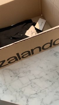 Zalando-kartonilaatikko, joka sisältää mustan napitettu paidan, huolto-ohjeet näkyvissä, lepäämään marmoripinnalla.