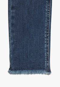 Jean en denim avec un ourlet frangé, de couleur bleu foncé, doté de coutures visibles et d'une texture douce. Le tissu est légèrement décoloré avec une finition lisse.