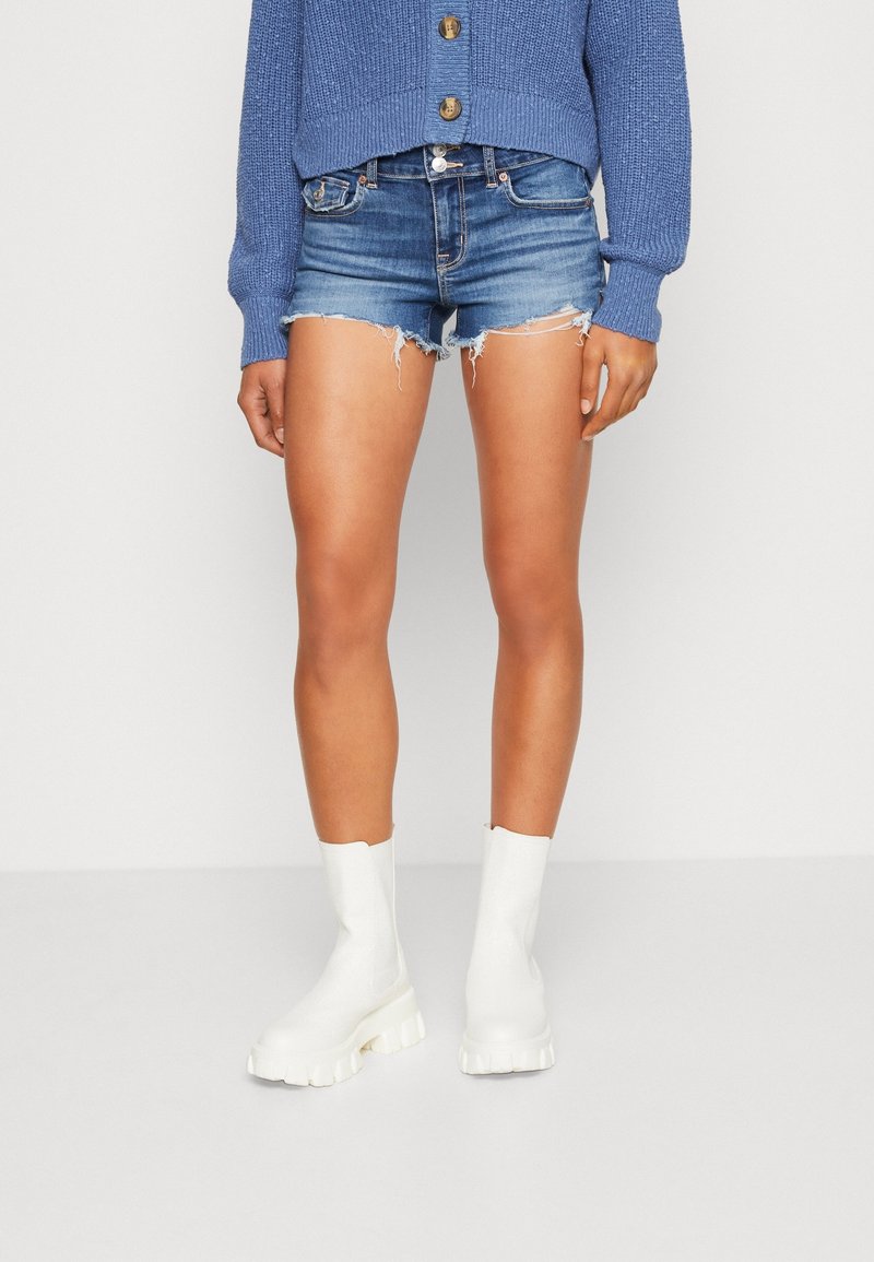 Femme portant un short en jean déchiré, des bottes blanches épaisses et un cardigan en tricot bleu avec de gros boutons, sur un fond uni.