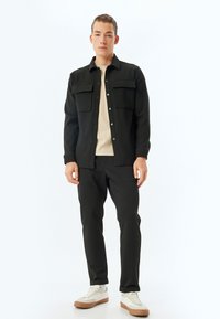 Giacca nera con bottoni, con due taschini sul petto; indossata sopra una maglietta beige a coste; abbinata a pantaloni neri e sneakers bianche con suole marroni.