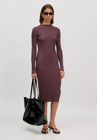Rippmuster, langärmliges Midi-Kleid in dunklem Mauve. Kombiniert mit einer glänzenden schwarzen Tote-Tasche. Schlichte Riemchensandalen vervollständigen den Look.