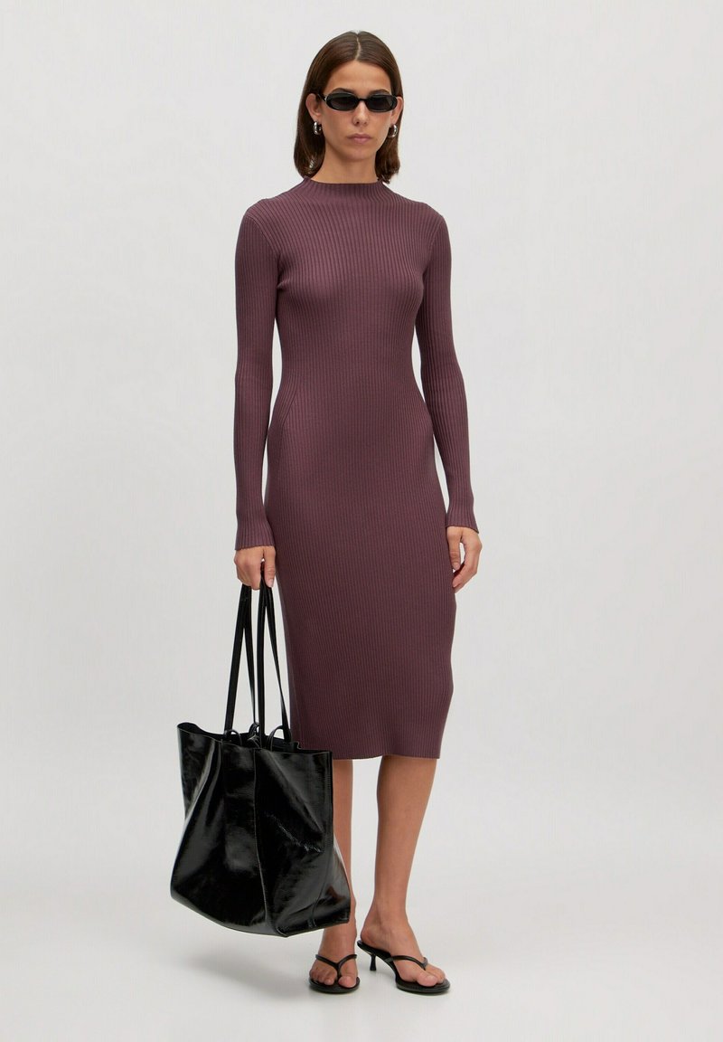 Rippmuster, langärmliges Midi-Kleid in dunklem Mauve. Kombiniert mit einer glänzenden schwarzen Tote-Tasche. Schlichte Riemchensandalen vervollständigen den Look.