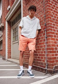 Gestreiftes kurzärmliges Hemd in Hellblau und Weiß, kombiniert mit korallenfarbenen Shorts und grünen Sneakers. Der Backsteinmauer-Hintergrund sorgt für Textur.