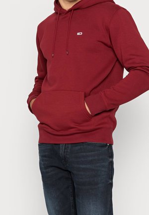 Mężczyzna ubrany w bordową bluzę z kapturem z przednią kieszenią i małym logo, w parze z ciemnoniebieskimi dżinsami, stoi z rękami w kieszeniach.
