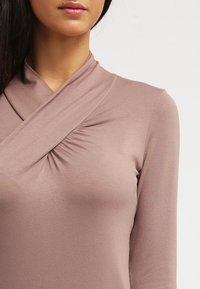 Frau trägt ein langärmliges, maulbeerfarbenes Wickeloberteil mit gerafftem Stoffdetail nahe der linken Schulter, schlichter Hintergrund.
