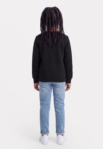 Dziecko z długimi skręconymi warkoczami, ubrane w czarną bluzę, jasnoniebieskie podwinięte jeansy i białe trampki, zwrócone tyłem na białym tle.