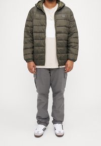 Olivgrön vadderad jacka med horisontell quiltning, huva och dragkedja fram; kombinerad med en beige t-shirt och grå cargobyxor. Vita sneakers.