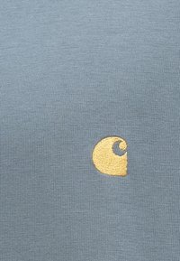 Κίτρινο κεντημένο λογότυπο Carhartt σε μπλε ριγωτό ύφασμα φόντο.