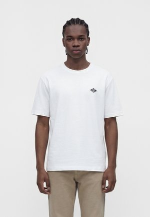 Jeune homme aux cheveux tressés portant un t-shirt blanc uni avec un petit logo sur la poitrine et un pantalon beige, debout devant un fond blanc.