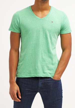 Homme portant un T-shirt vert clair à col en V et un jean bleu foncé, une main dans la poche et l'autre détendue sur le côté.