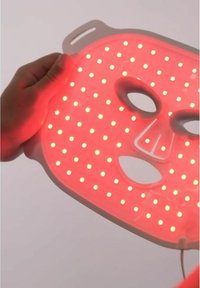 Masque facial LED rouge avec une surface lisse, de forme rectangulaire et plusieurs petits points lumineux. Découpes pour les yeux et la bouche, tenu par une main.