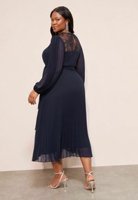 Tumes pleediga kleit pika varrukaga, pitsist seljatükk, figuuri rõhutav vöökoht ja pahkluuni ulatuv seelik; koos hõbedaste rihmadega kontsadega.