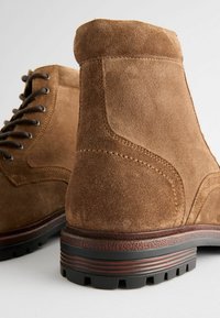 Cizmele de gleznă din suede maro au vârf rotund, design cu șireturi, cusături contrastante și o talpă din cauciuc texturată cu caneluri.
