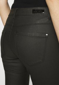 Pantalones de mezclilla negra con un diseño elegante, textura suave y dos bolsillos traseros con herrajes plateados. Incluye una etiqueta de marca en la cintura.