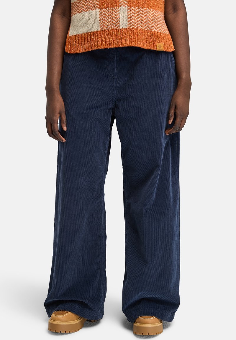 Personne portant un pantalon large en velours côtelé bleu marine, des chaussures compensées marron clair et un pull orange à motifs coupé à la taille.