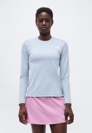 Kobieta ubrana w jasnoniebieską koszulę z długim rękawem i różową spódnicę, stojąca, zwrócona przodem na jednolitym tle.