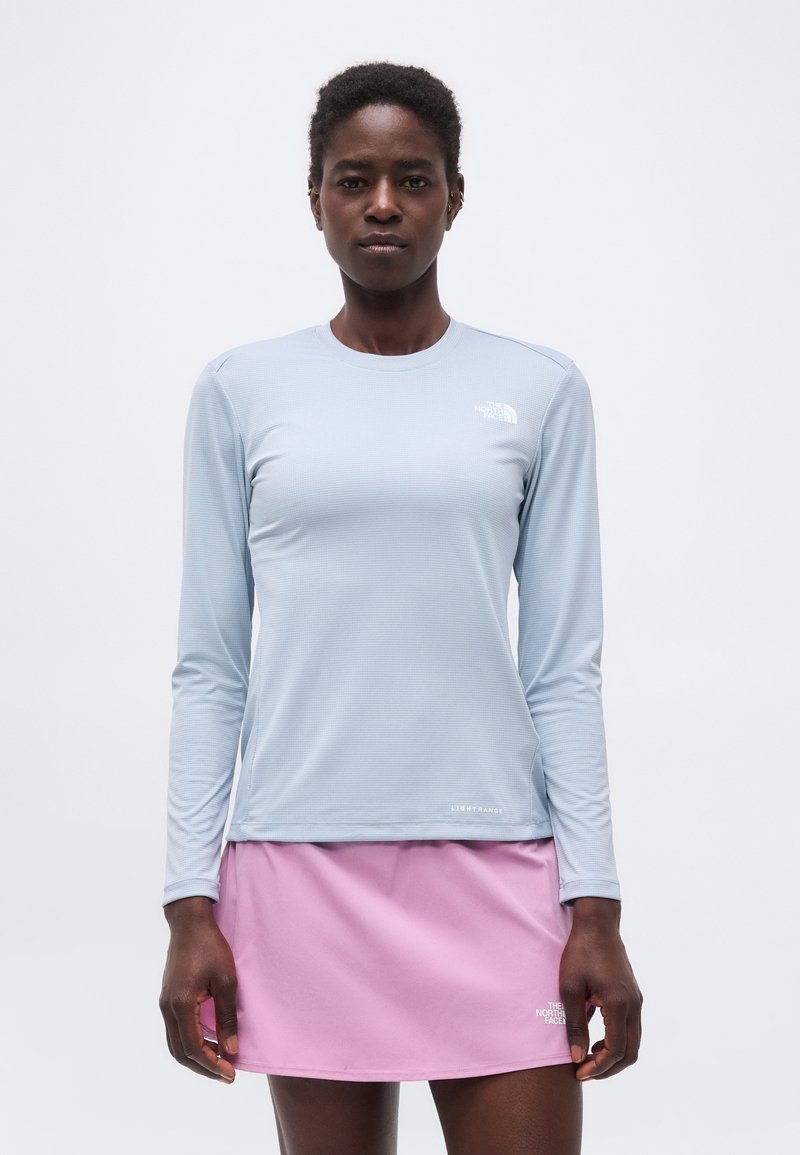 Γυναίκα που φοράει γαλάζιο πουκάμισο με μακριά μανίκια και ροζ φούστα, όρθια, κοιτάζοντας μπροστά με φόντο απλό φόντο.