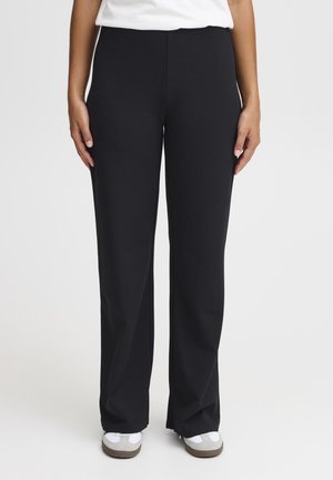 Une femme se tient debout vêtue d'un pantalon noir à jambes droites, de baskets blanches et d'un haut blanc rentré, sur un fond uni.