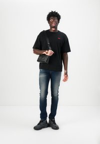 T-shirt noir à manches courtes avec logo rouge, jean en denim bleu, sac bandoulière noir et chaussures de sport noires avec un design texturé.