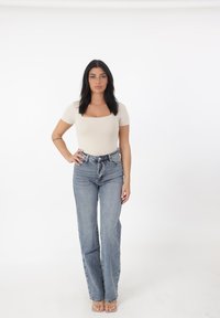 Top beige ajustado, jeans de tiro alto de color azul claro y sandalias transparentes. Diseño simple con acentos mínimos, de pie contra un fondo blanco liso.