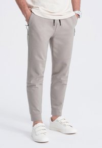 Pantalones de chándal gris claro en tejido suave, con puños elásticos, bolsillos laterales y detalles de cintura con cordón. Combinados con zapatillas blancas sin cordones.