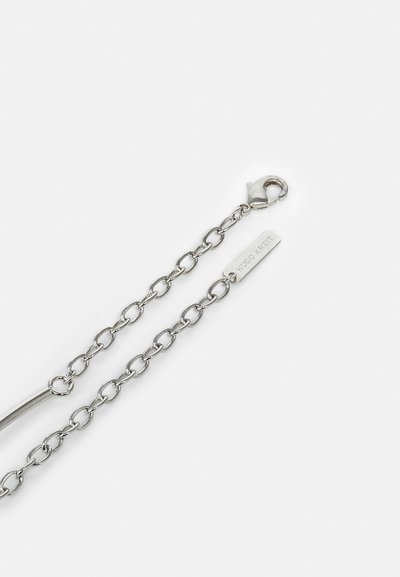 Catena in tono argento con anelli allungati, dotata di chiusura a moschettone e un tag piatto inciso con "HUGO KREIT." Texture liscia e finitura metallica.