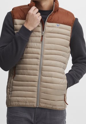 Mężczyzna zapina beżowo-brązową pikowaną kamizelkę na ciemną, długą koszulę, jedną ręką przy kołnierzu, drugą w kieszeni kamizelki.