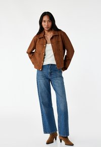 Ruskea mokkatakkia, jossa on kauluskotelo, etupuolella napit, ja kaksi sivutaskua. Yhdistetty korkeavyötäröisiin sinisiin farkkuihin.