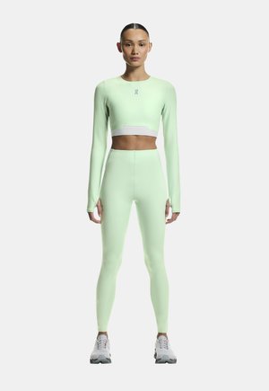Kvinde stående iført lysegrøn langærmet crop top og matchende højtaljede leggings med hvide sportssko på hvid baggrund.