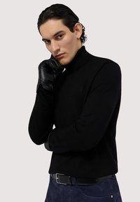 Svart turtleneck-tröja med ribbad struktur, kombinerad med svarta läderhandskar. Diskret logotypdetalj på bröstet, bärs med mörkblå byxor.