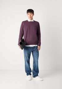Lilla hettegenser med en subtil logo, hvit skjorte med krage under, blå jeans og hvite joggesko, holder en svart veske i den ene hånden.