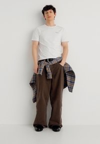 Νεαρός άνδρας που στέκεται με τα χέρια στις τσέπες, φοράει λευκό μπλουζάκι, καφέ παντελόνι φαρδύ στο κάτω μέρος, μαύρα παπούτσια και πουκάμισο καρό δεμένο στη μέση.