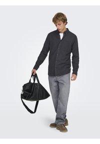 Langærmet mørkegrå skjorte med knapper og brystlomme, lysegrå jeans og brune ruskindsstøvler, der holder en stor sort læderduffeltaske.